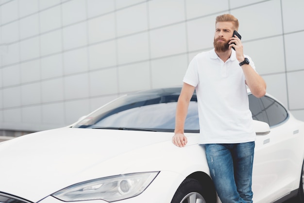 Cara com barba falando no telefone perto de seu carro elétrico.