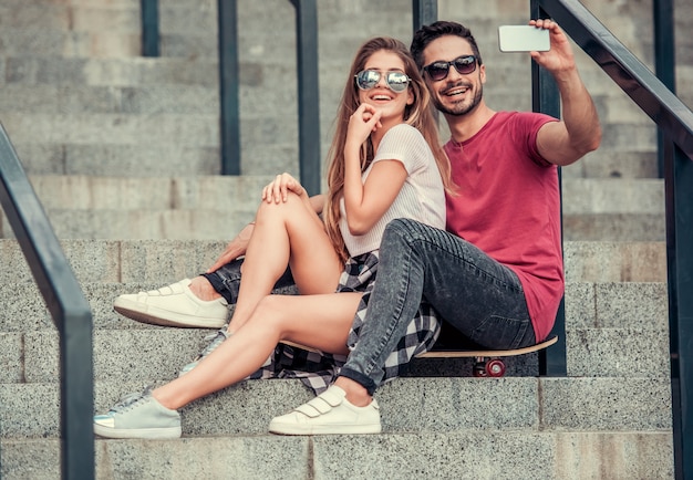 cara com garota nos degraus leve selfie no telefone e sorria. Jovem casal tendo selfie telefone enquanto está sentado no skate.