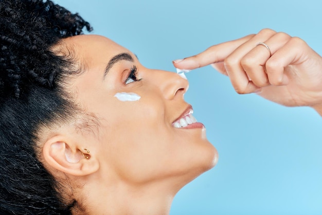 Foto cara de cuidado de la piel de belleza y mujer feliz con aplicación de crema de loción humectante o producto para la prevención de espinillas maquillaje de bienestar cosméticos antienvejecimiento o perfil de persona de estudio sobre fondo azul