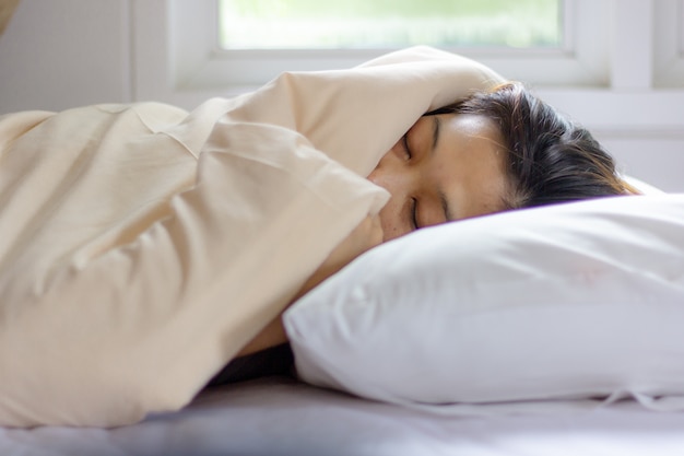 Cara da jovem mulher que dorme ao encontrar-se na cama branca no alvorecer do raio de sol.