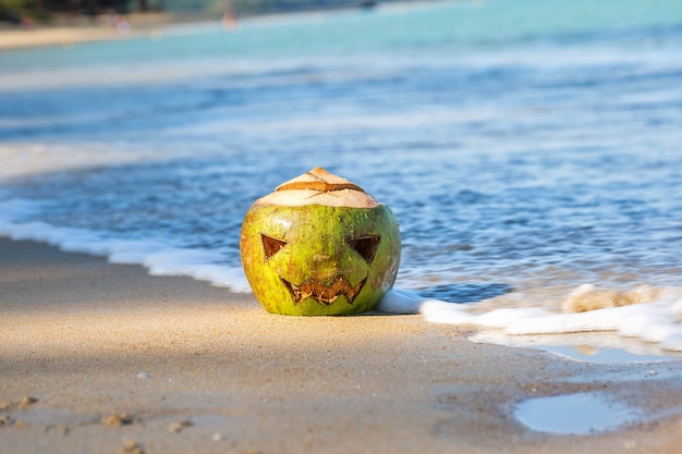 Cara de coco verde esculpida como abóbora de halloween deita na areia na praia tropical jack o lantern cabana