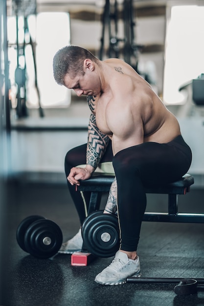 Cara de fisiculturista motivado se senta em um banco e realiza exercícios para seus bíceps.