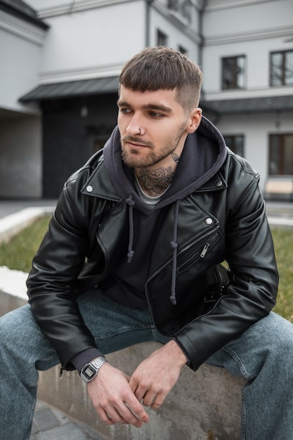 Cara de hipster elegante bonito na moda com cabelo barba e tatuagem no pescoço em roupa casual com casacos com capuz e jeans senta-se na cidade Homem modelo na moda