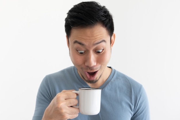 La cara divertida del hombre asiático en camiseta azul bebe café de la taza blanca.