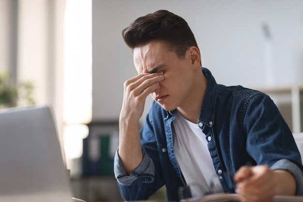 Cara do milênio cansado que sofre de fadiga ocular ao usar o laptop no escritório