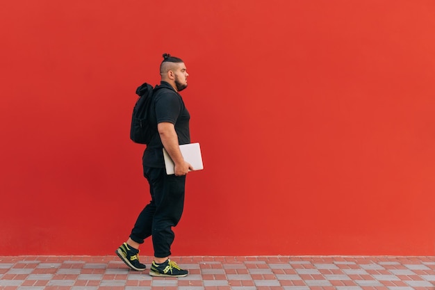 Cara elegante com excesso de peso com barba usa roupas elegantes caminha no contexto de uma parede vermelha