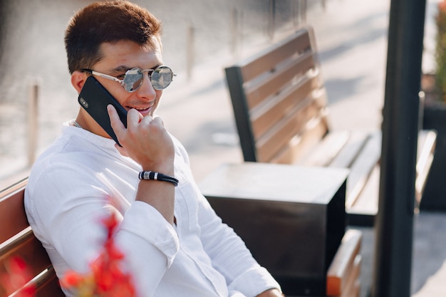 Cara elegante jovem de camisa com telefone no banco em telefonemas ensolaradas de dia quente ao ar livre