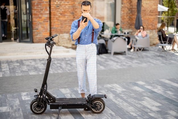 Cara elegante usa telefone enquanto está perto de sua scooter elétrica ao ar livre