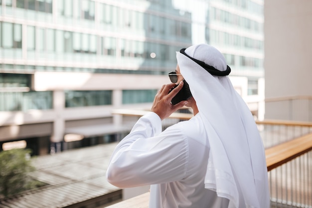 Cara emirati vestindo kandura tradicional no estilo de vida urbano da cidade emirados, falando com telefone inteligente.