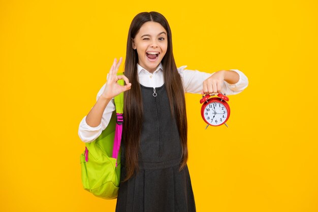 Cara engraçada De volta à escola A menina da escola adolescente com mochila segura a hora do despertador para aprender Crianças em idade escolar em fundo amarelo isolado