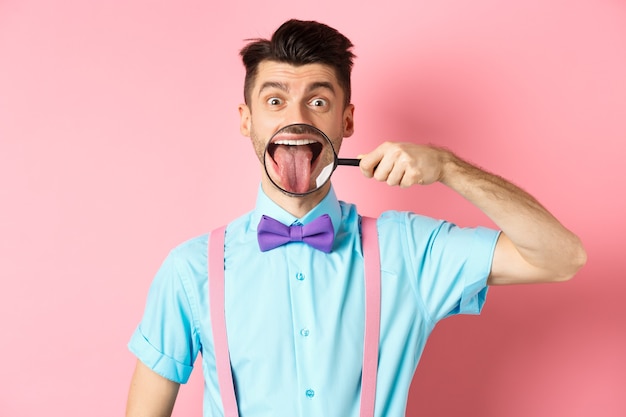 Cara engraçado caucasiano mostrando a língua na lupa, sorrindo e fazendo caretas, de pé na gravata borboleta sobre fundo rosa.