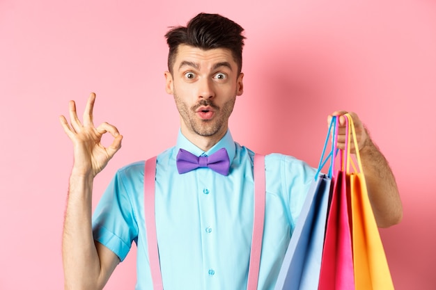 Cara engraçado em gravata borboleta mostrando sinal de OK e sacolas de compras, elogiando um bom negócio nas lojas, em pé sobre rosa.