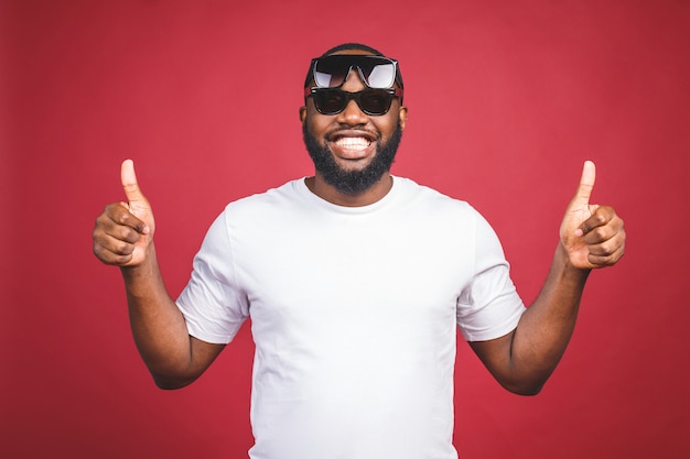 Cara engraçado em t-shirt branca e sol glases pulando e olhando para a câmera. retrato do estúdio do modelo masculino africano emocional que levanta no fundo vermelho. afirmativo.