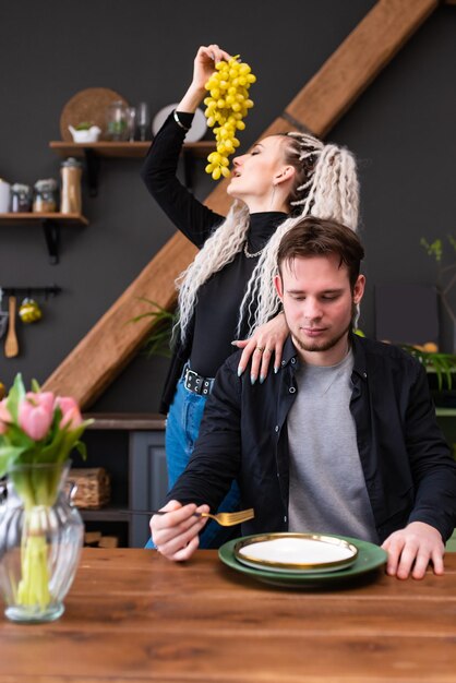 Cara está sentado à mesa com um garfo na mão na frente de um prato vazio Atrás da garota come uvas Jovem casal alegre em roupas casuais