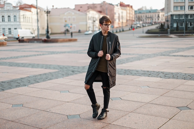 Cara estiloso com óculos em uma jaqueta da moda e jeans rasgados com tênis preto na cidade