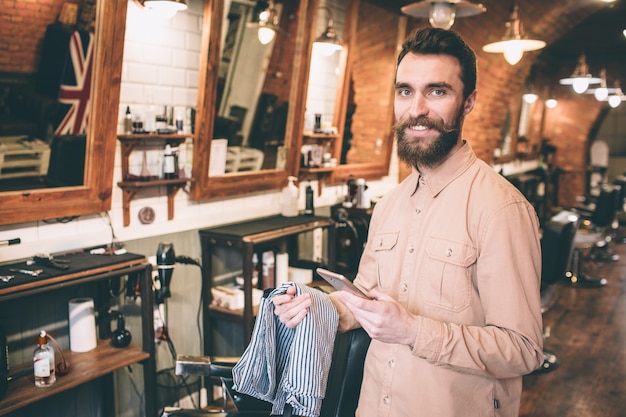 Cara feliz e alegre está de pé em uma barbearia e olhando para a câmera. Ele está sorrindo. Homem está segurando uma coisa de capa para os ombros do cliente.