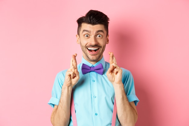 Cara feliz fazendo um desejo, segurando os dedos cruzados para dar sorte e sorrindo para a câmera, esperando uma boa notícia, em pé sobre um fundo rosa.