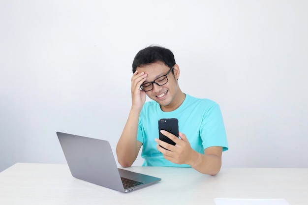 Cara feliz de un joven asiático cuando hace una videollamada o qué en el teléfono con una computadora portátil junto a un fondo gris aislado Hombre indonesio con camisa azul