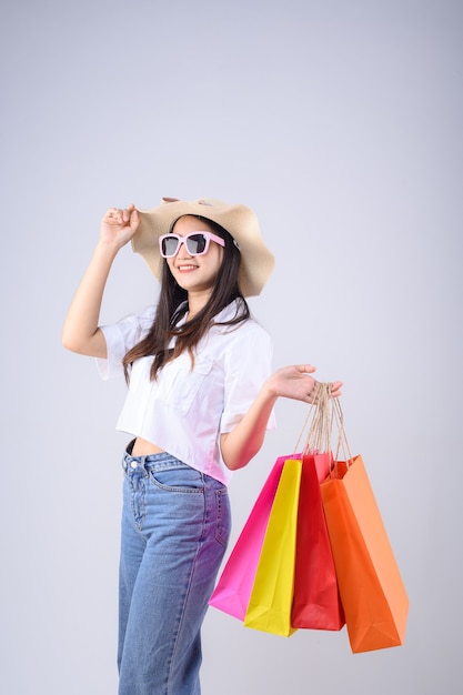 Foto cara feliz joven mujer asiática sosteniendo una bolsa de compras, con sombrero y gafas aisladas sobre fondo blanco.