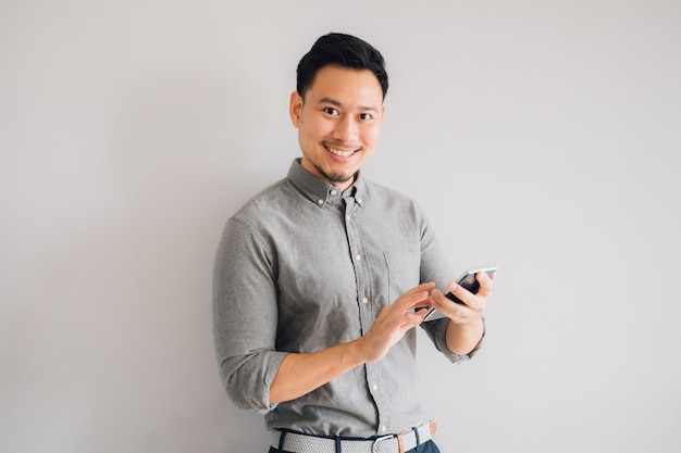 Cara feliz de la sonrisa del soporte asiático hermoso del smartphone del uso del hombre aislado en fondo gris.