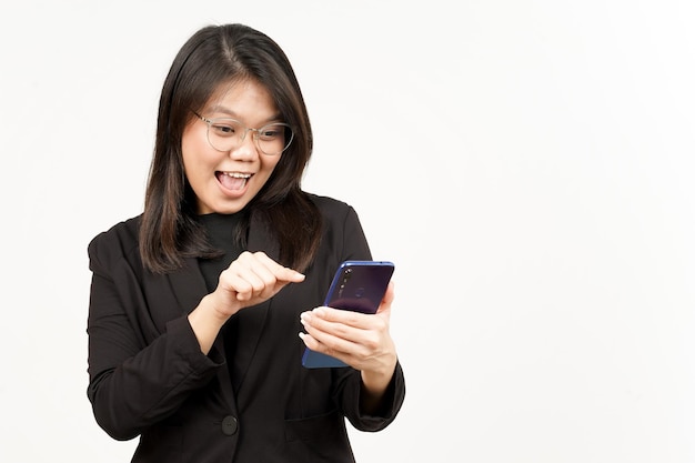 Cara feliz y usando el teléfono inteligente de una hermosa mujer asiática con chaqueta negra aislada en blanco