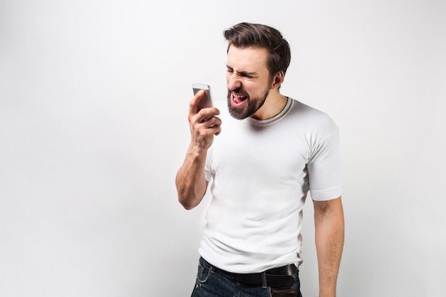 Cara forte e grande está falando ao telefone em voz alta. Ele está gritando palavras que saem de sua boca. Jovem está expressando emoções ruins. Isolado na parede branca