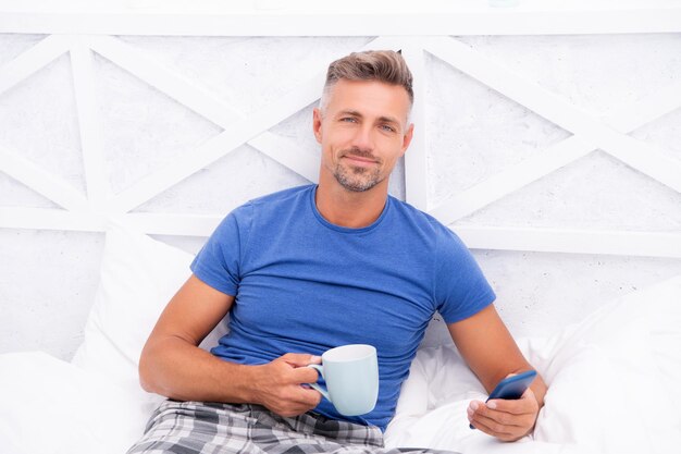 Cara grisalho lendo notícias matinais no celular enquanto toma café na cama