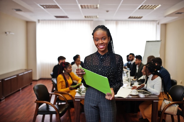 Cara de una hermosa mujer de negocios africana sosteniendo un portapapeles en el fondo de una reunión de equipo multirracial de gente de negocios sentada en una mesa de oficina