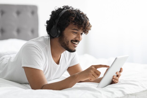 Cara hindu relaxado usando tablet digital e fones de ouvido na cama