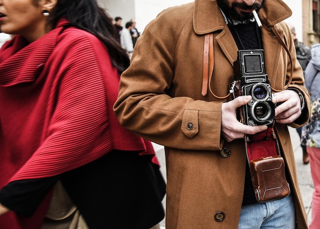Cara hipster com a câmera vintage fotografando pessoas na cidade Fotojornalista com uma famosa câmera retrô tirando foto no meio da multidão durante demonstração de rua Estilo de fotografia de rua