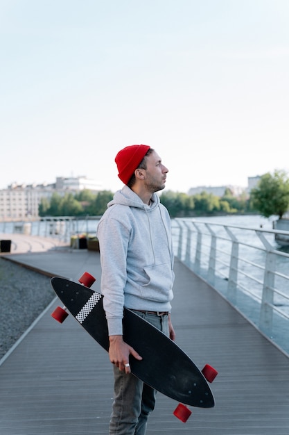 Cara hipster com longboard na ponte da cidade. Skatista moderno segurando o skate na mão