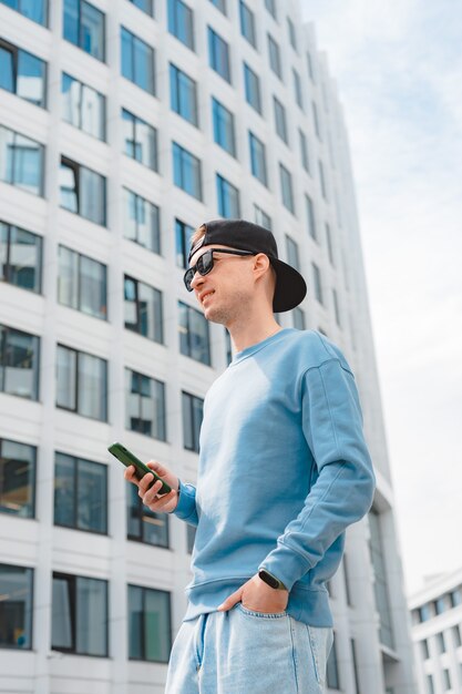 Cara hipster positivo com mochila em pé ao ar livre segurando o smartphone enquanto desvia o olhar com um sorriso