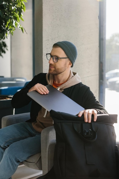 Cara jovem bonito freelancer trabalhando com laptop no escritório Um homem de óculos e um chapéu está segurando um laptop enquanto está sentado em uma cadeira no corredor