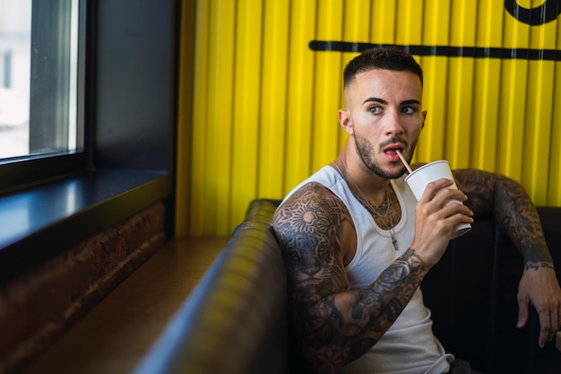 Cara jovem caucasiano com tatuagens legais sentado no café e bebendo um copo de suco