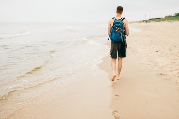 Foto cara jovem com mochila atrás vai com os pés descalços na praia