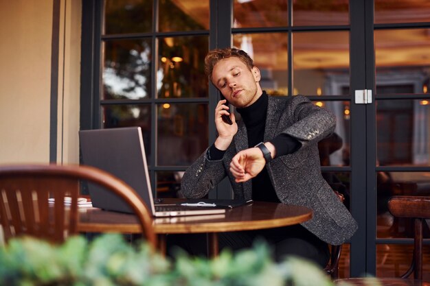 Cara jovem elegante com roupa formal se senta no café com seu laptop e fala ao telefone.