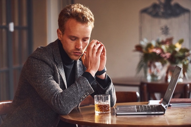 Cara jovem elegante com roupa formal se senta no café com seu laptop e um copo de álcool.