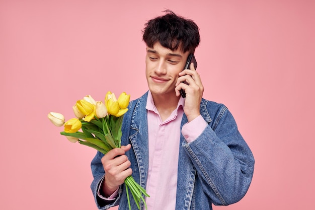 Cara jovem em uma jaqueta jeans com um buquê de flores falando ao telefone fundo rosa