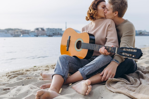 Cara jovem ensinando sua namorada a tocar violão autodidata Casal apaixonado se divertindo na praia abraçando e beijando