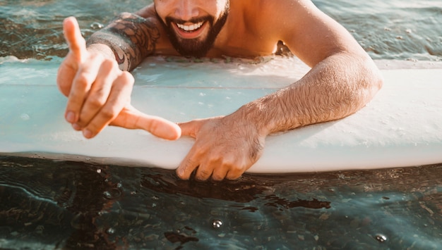 Cara jovem feliz com gesto shaka deitado na prancha de surf na água