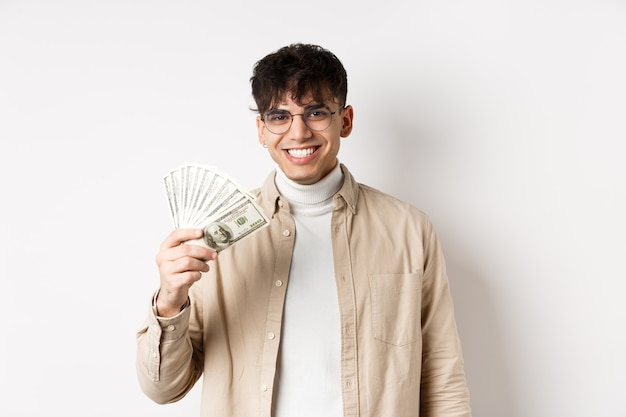 Cara jovem feliz segurando notas de dólar e sorrindo, ganhando dinheiro e de pé na parede branca.