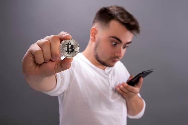 Cara jovem fica surpreso ao assistir o mercado de criptomoedas em seu telefone segurando uma moeda bitcoin na mão