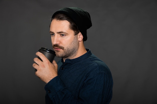 Foto cara jovem hippie com chapéu de malha tomando café em fundo escuro