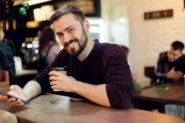 Cara jovem hippie enviando mensagens de texto com seu celular no bar e tomando um cappuccino hipster de homem da moda jovem bebendo café expresso no café da cidade durante a hora do almoço e trabalhando no computador tablet