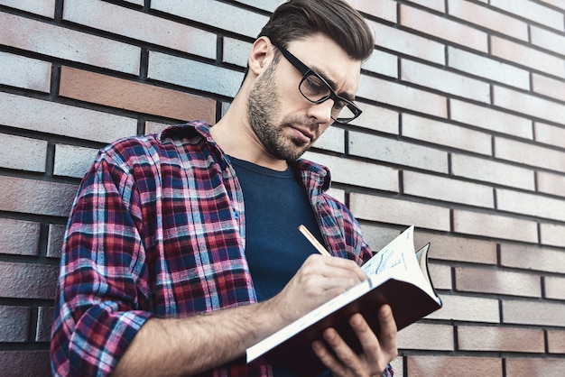 Foto cara jovem hippie usando óculos, lendo um livro ou caderno, fazer algumas anotações e idéias sobre o fundo da parede de tijolo.