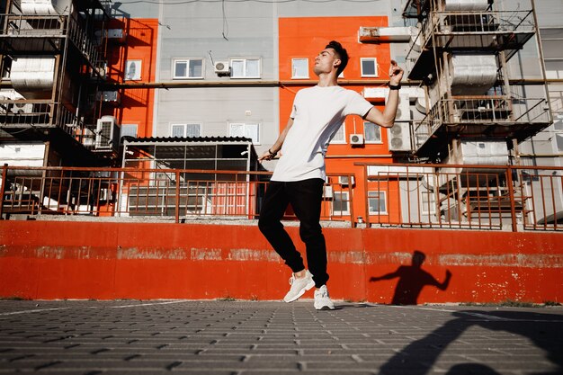 Cara jovem vestido de jeans e camiseta está dançando a dança moderna na rua ao fundo dos edifícios urbanos no dia quente.