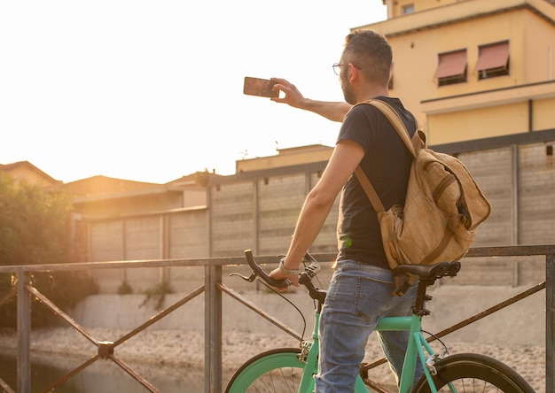 Foto cara jovem visto por trás de pé em sua bicicleta minimalista usando sua mochila faz uma selfie com seu smartphone ao pôr do sol