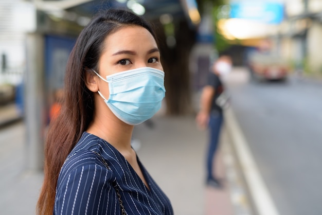 Cara de joven empresaria asiática con máscara para protegerse del brote del virus corona esperando en la parada de autobús