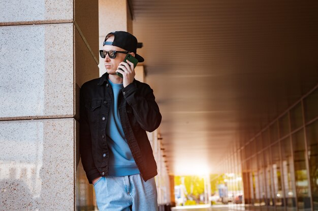 Cara milenar hippie em roupa elegante, em pé ao ar livre, falando em um smartphone