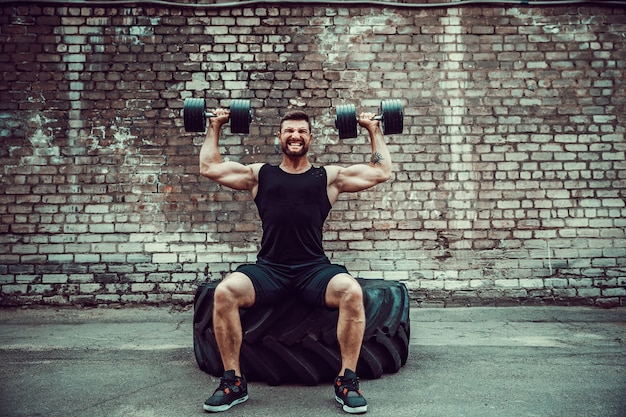 Cara musculoso fazendo exercícios com halteres contra uma parede de tijolos